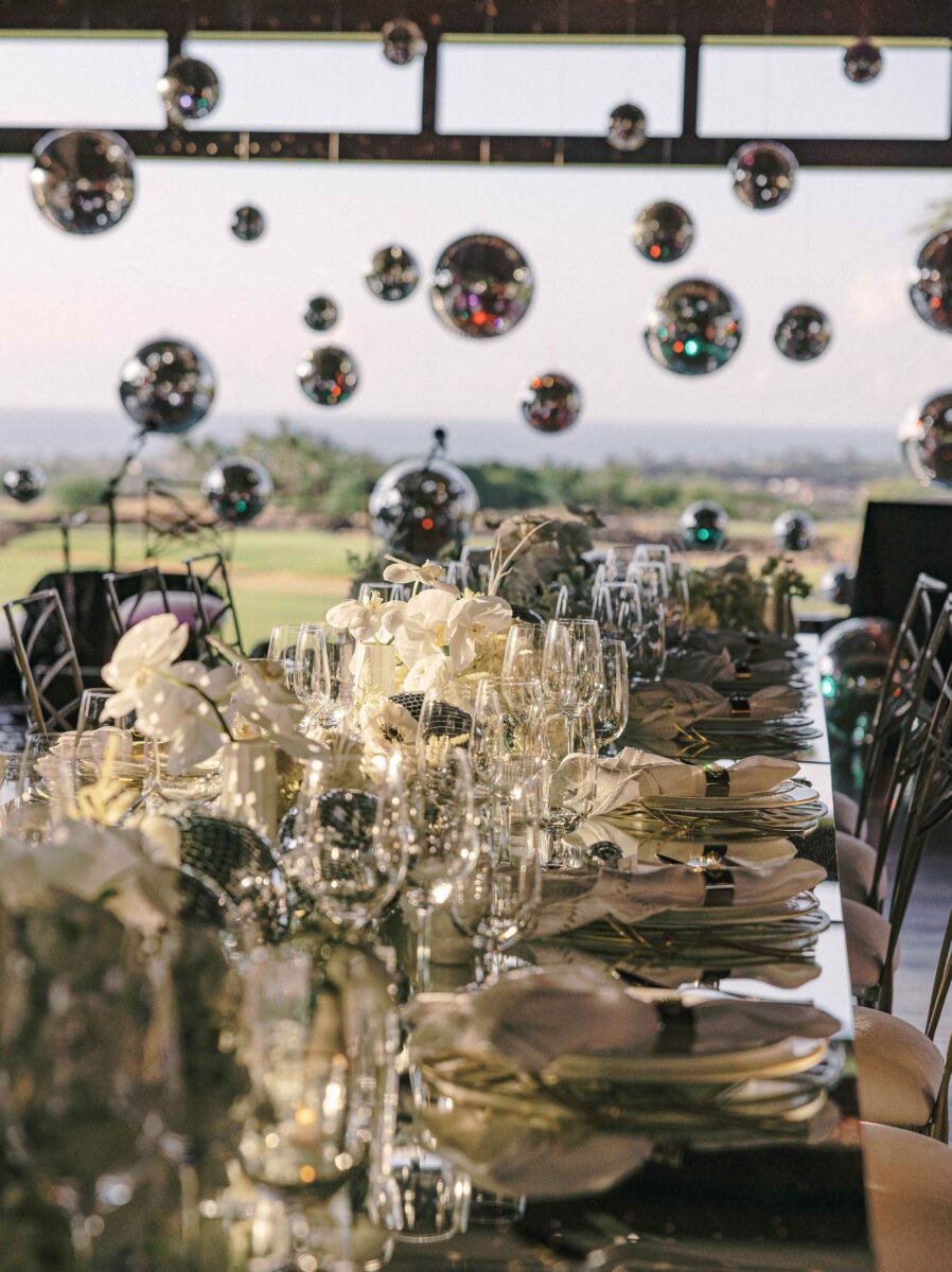 Elegantly set table with assorted glassware and dishes, white flowers, and 20 disco balls hanging in background. 