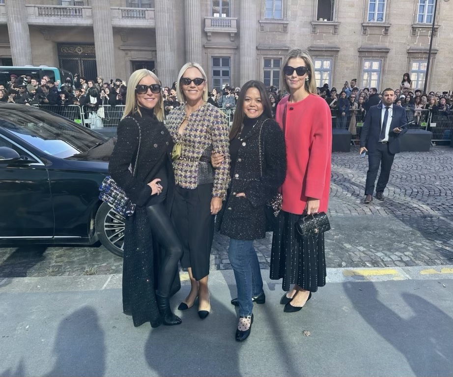 Four women dressed elegantly standing on streets of Paris in front of crowd awaiting fashion show