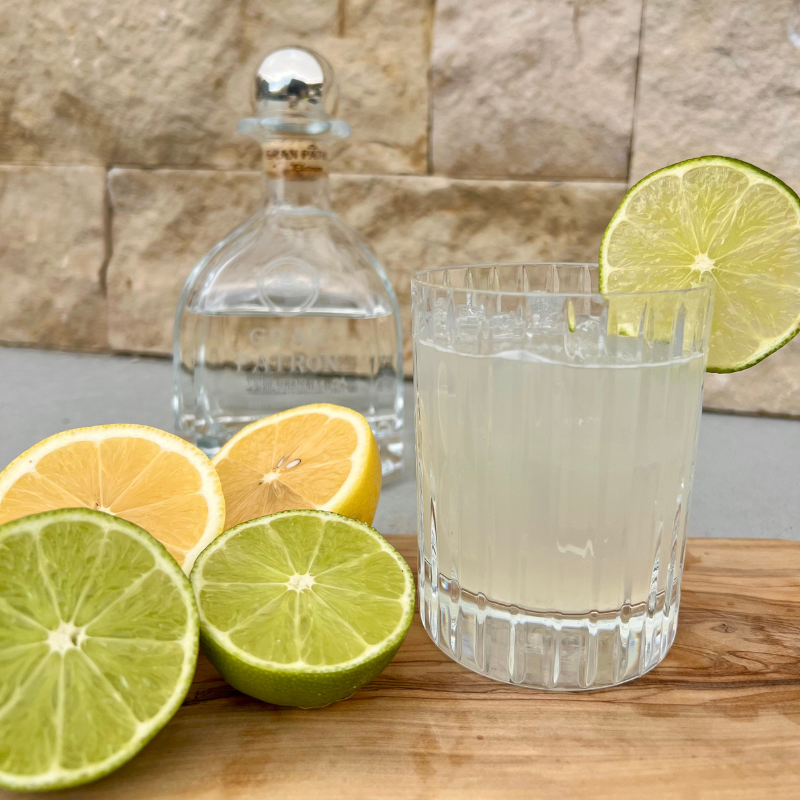 Margarita over ice garnished with lime wheel sitting on wood cutting board next to cut lemons and limes and bottle of Patron Platinum