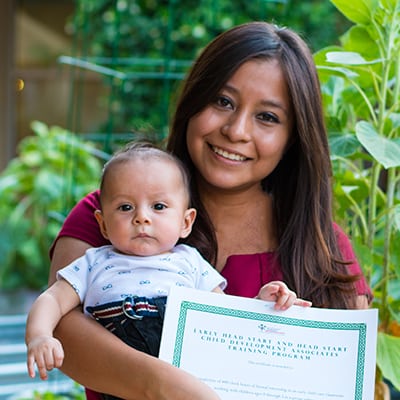 Woman holds her baby in her left arm and holds a certificate in her right hand
