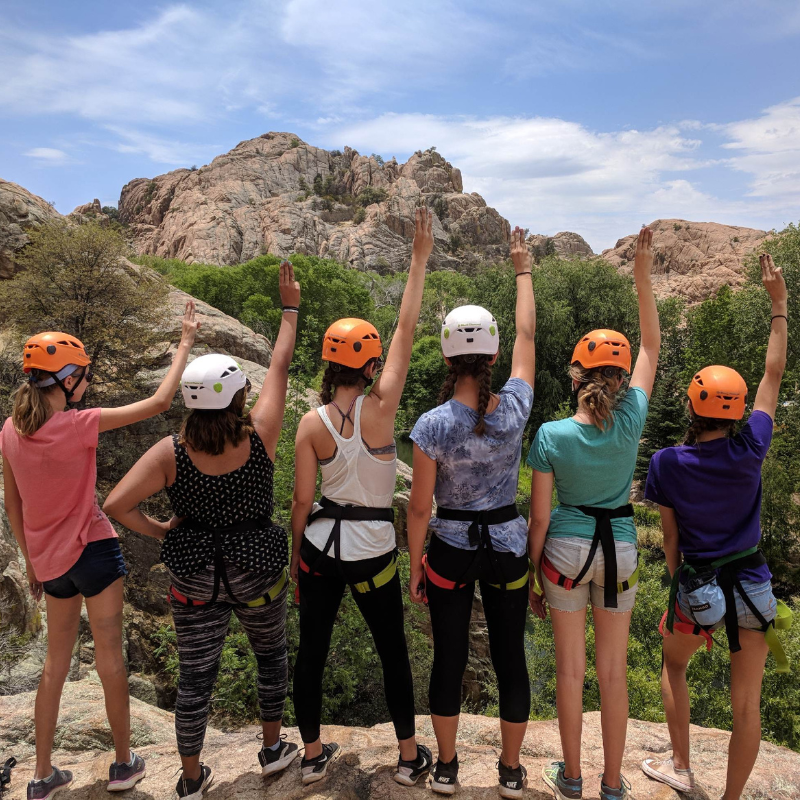 Young girls in helmets and rock climbing harnesses stand on a rock looking out towards the mountains 