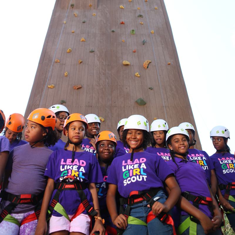 Young girls in helmets and rock climbing harnesses smile in front of a rock wall